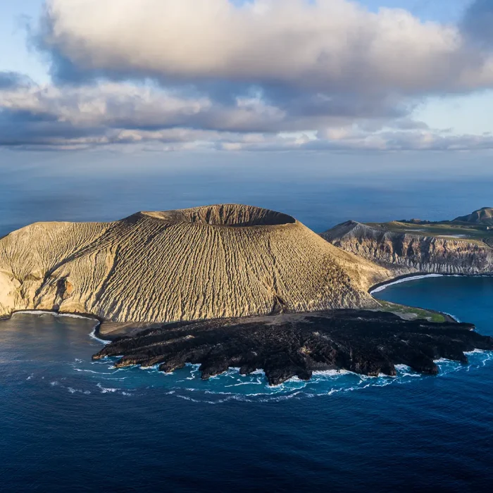 Aerial view of Cocos Island National Park, featuring a volcanic island with a large crater, rugged slopes, and dark rocky shoreline, surrounded by blue ocean under a partly cloudy sky.