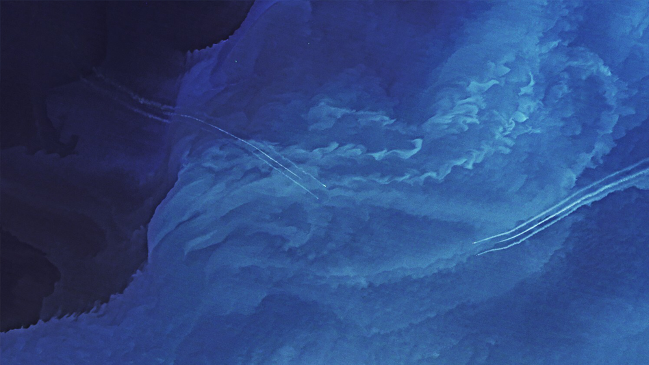 Aerial view of swirling blue patterns resembling ocean waves or clouds, with several thin white contrails crossing through the center.