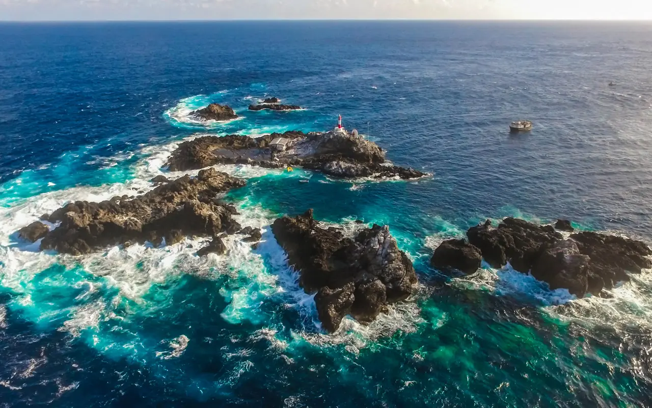 Aerial view of rocky islets surrounded by turquoise ocean water and white waves, with a small red lighthouse perched on one of the rocks under a bright sky, captured using high resolution satellite imagery.