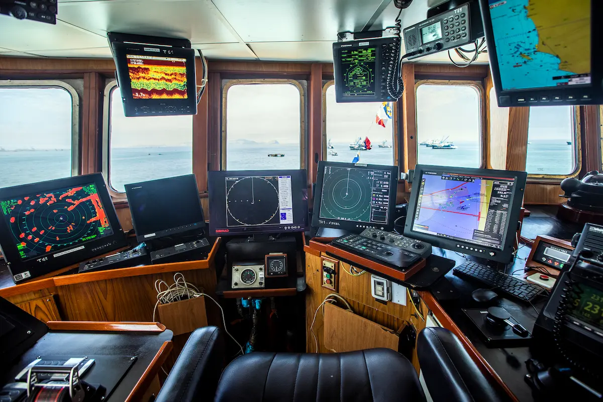 La imagen muestra la sala de control de un barco, con múltiples pantallas de radar y navegación, paneles de control y grandes ventanales con vistas al mar y a los barcos que se divisan en la lejanía.