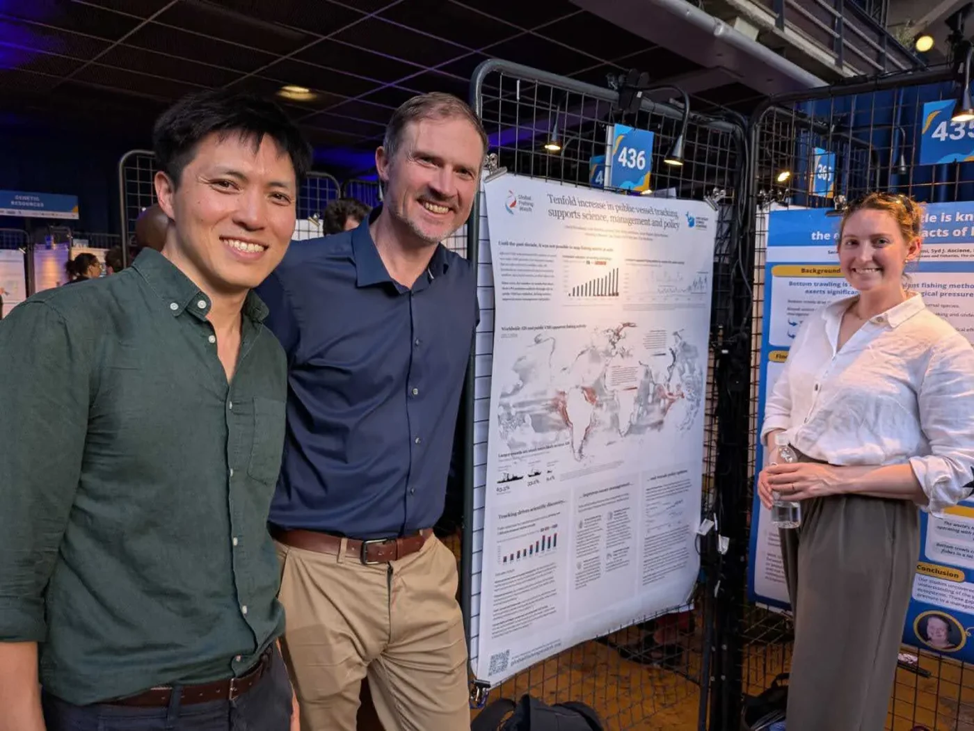 Trois personnes sourient devant un poster de recherche scientifique comportant des graphiques et une carte du monde, lors d'un événement en intérieur, avec d'autres posters en arrière-plan.
