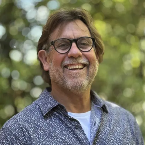 A middle-aged man with glasses, gray hair, and a beard smiles outdoors, wearing a patterned shirt over a white t-shirt. The background is blurred greenery with sunlight.