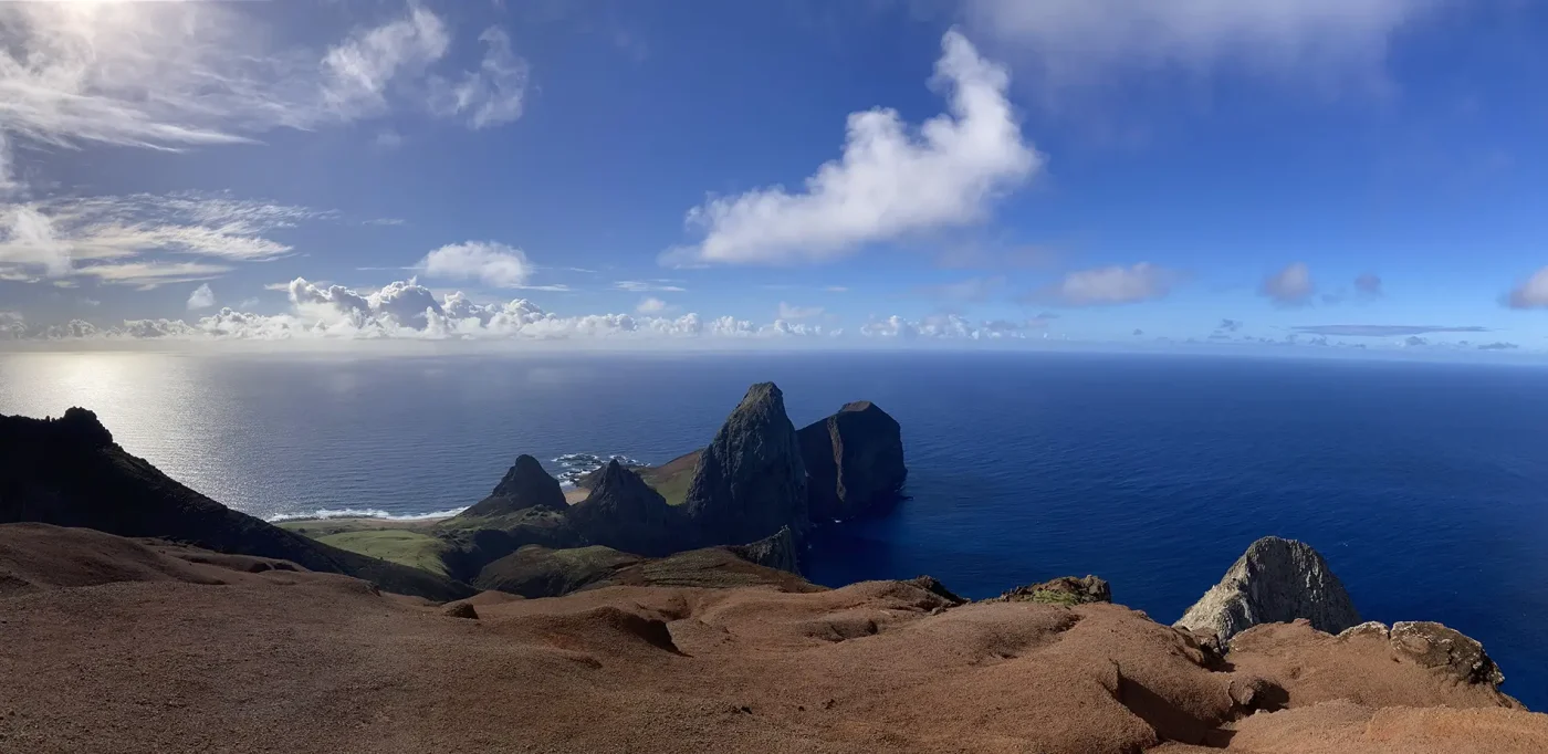 Panoramic view of rugged coastal cliffs and rocky islands, captured with high-resolution satellite imagery, shows the deep blue ocean under a bright sky. Brown, rocky foreground contrasts with the vibrant sea and sky.
