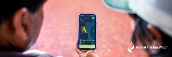 Two people look at a smartphone displaying a map with highlighted fishing activity. The Global Fishing Watch logo appears in the bottom right corner of the image.