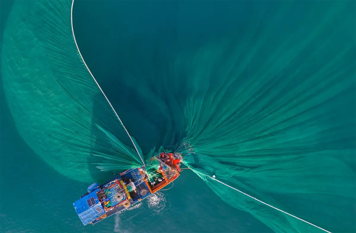 Aerial view of a fishing boat pulling large green nets through clear blue water, creating sweeping patterns on the waters surface.