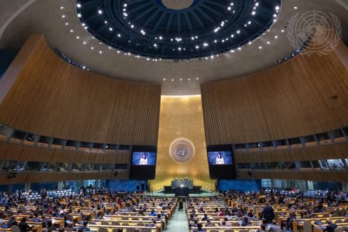 La sala de la Asamblea General de las Naciones Unidas, llena de delegados sentados en pupitres, está frente a un escenario central con un podio y un gran emblema de la ONU en una pared dorada; dos pantallas muestran a un orador que se dirige a la asamblea.