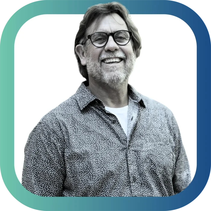A smiling middle-aged man with glasses, a beard, and a patterned shirt stands against a white background, framed by a rounded border with a blue and green gradient.