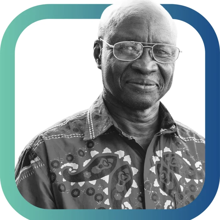Black and white photo of an older man wearing glasses and a patterned shirt, smiling slightly. The image is framed by a rounded teal and blue border.