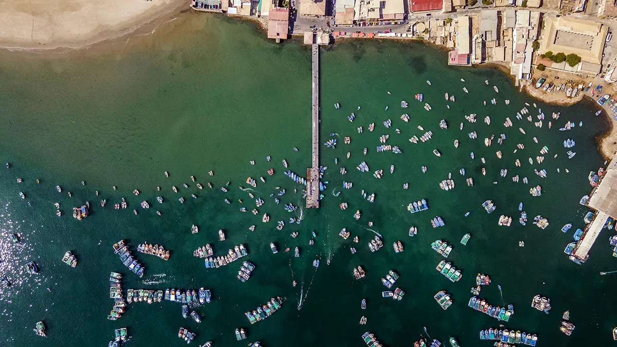 Aerial top-down view of a pier stretching into emerald green waters crowded with dozens of small fishing boats near a coastal town and sandy beach.