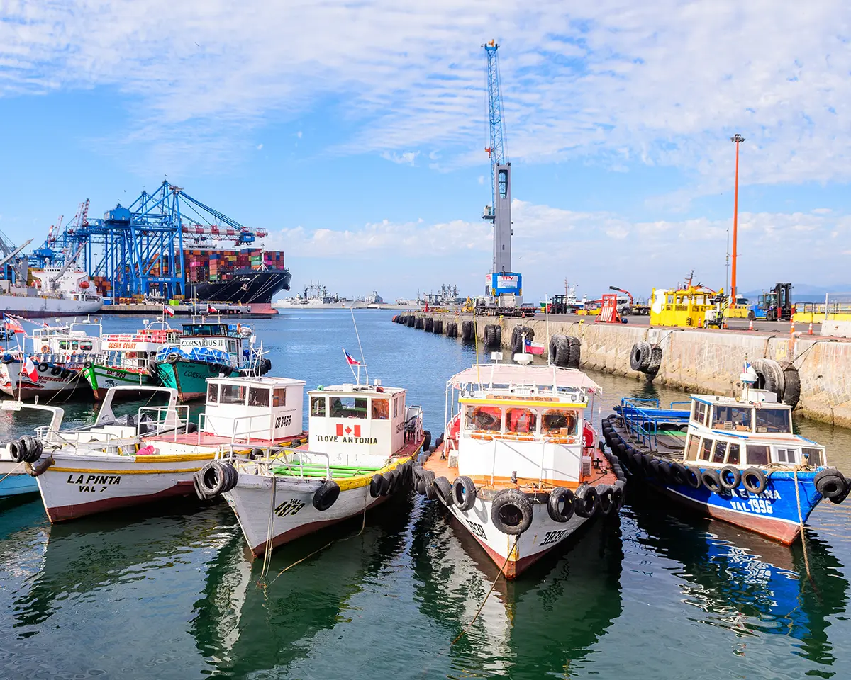 Several small fishing boats are docked side by side in a busy harbor, with cranes, large ships, and industrial equipment visible in the background under a partly cloudy sky.