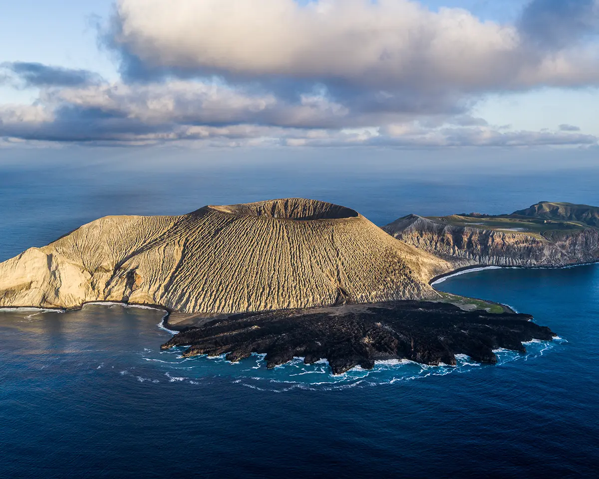 A rugged volcanic island with steep, eroded slopes rises from deep blue ocean waters, bordered by white-capped waves and under a partly cloudy sky.