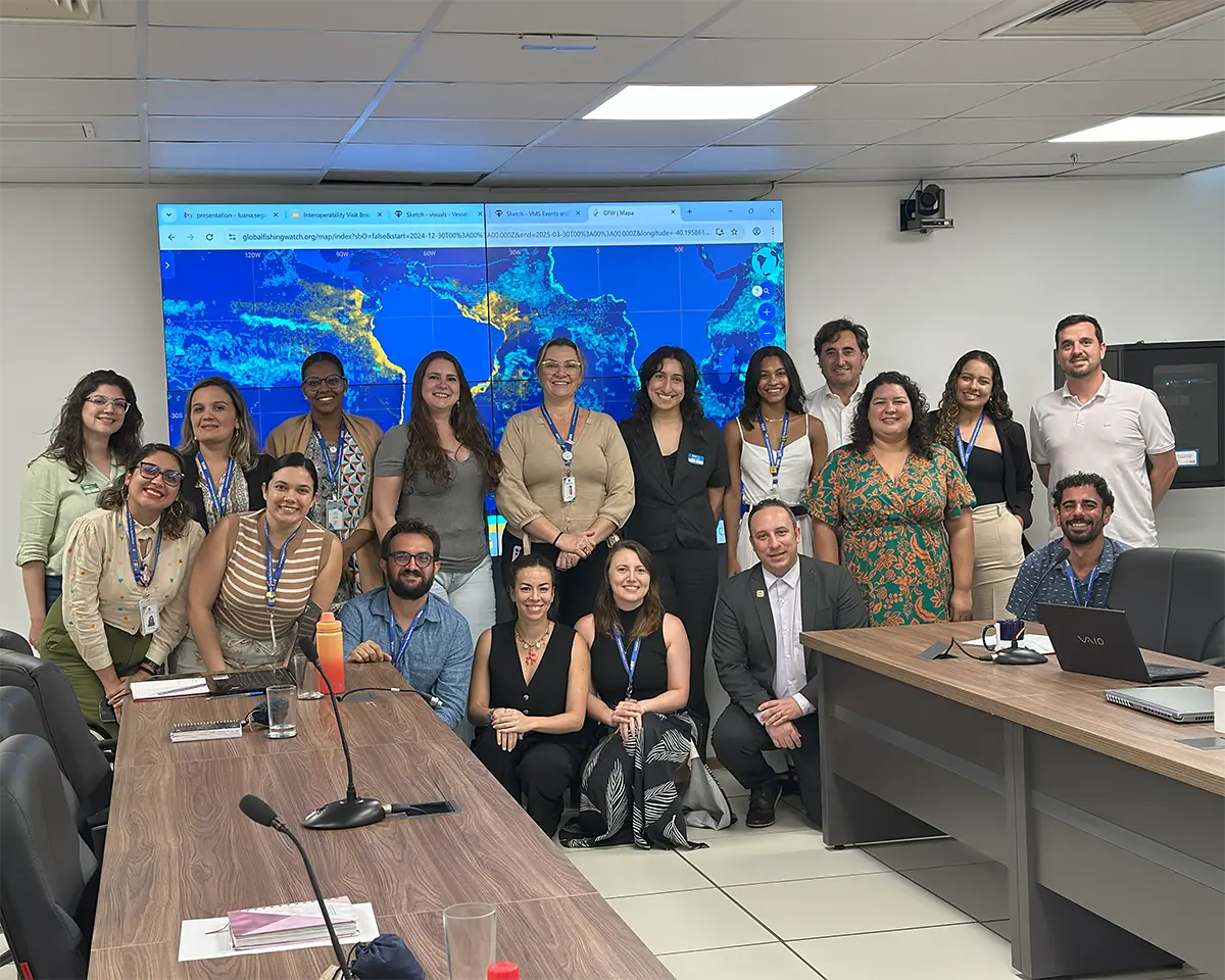 A group of 20 people pose and smile together in a conference room, with some seated and others standing. Behind them is a large screen displaying a colorful map. Microphones and papers are on the tables in front.