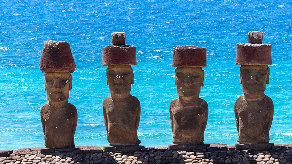 Four Moai statues with red stone hats stand in a row on a stone platform, with bright blue ocean water sparkling in the background under sunlight.