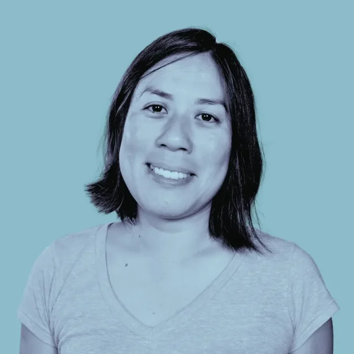 A woman with shoulder-length dark hair smiles at the camera. She wears a light V-neck shirt, with a solid light blue background and blue-toned filter.