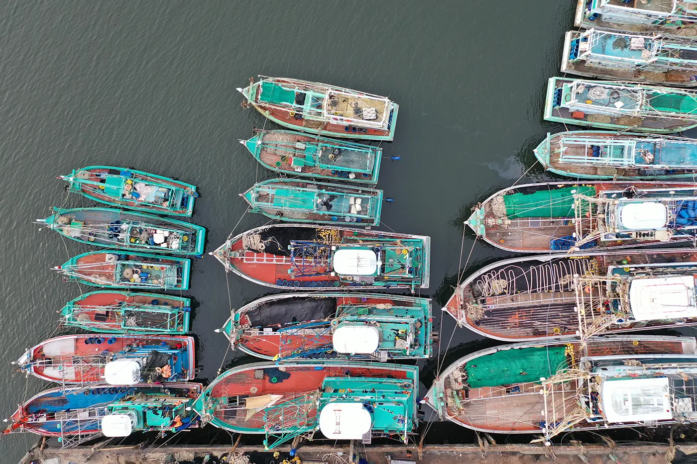 Vista aérea de coloridos barcos de pesca atracados uno junto al otro en un muelle, flotando en aguas tranquilas y oscuras.