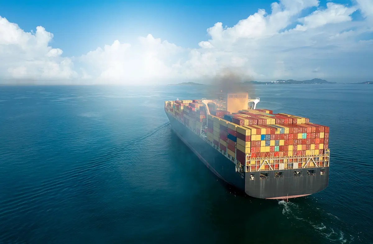 A large cargo ship loaded with colorful shipping containers sails on calm blue water under a bright, partly cloudy sky, with a trail of smoke rising from its smokestack.