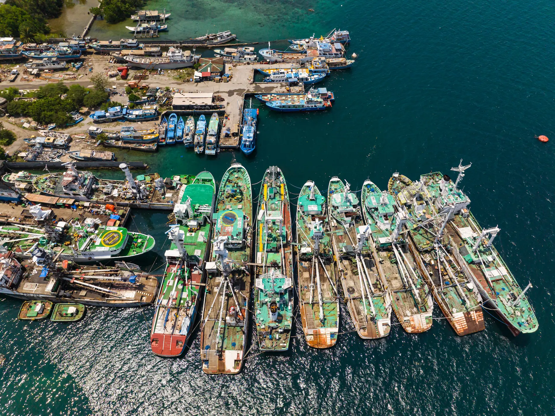 Aerial view of multiple fishing boats docked closely together at a busy harbor within Cocos Island National Park, with more boats and equipment visible on the shore nearby.