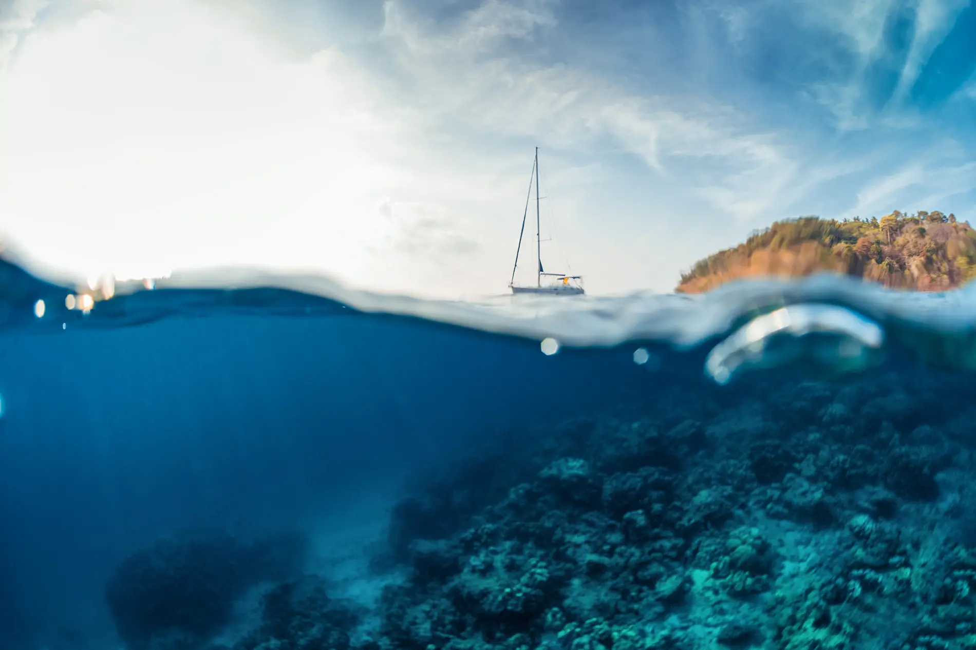 Split view of a sailboat on the ocean, with the top half showing sky and distant coastline, and the bottom half revealing underwater coral reefs through remarkable ocean transparency and clear blue water.