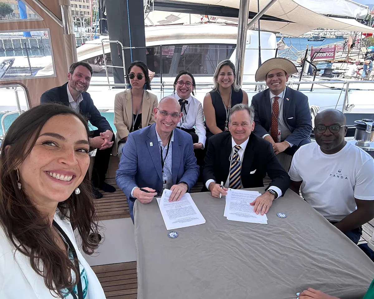 Un groupe de neuf personnes, vêtues de tenues professionnelles et décontractées, sourit pour une photo autour d'une table avec des documents sur un yacht, avec un port de plaisance et des bateaux visibles à l'arrière-plan.