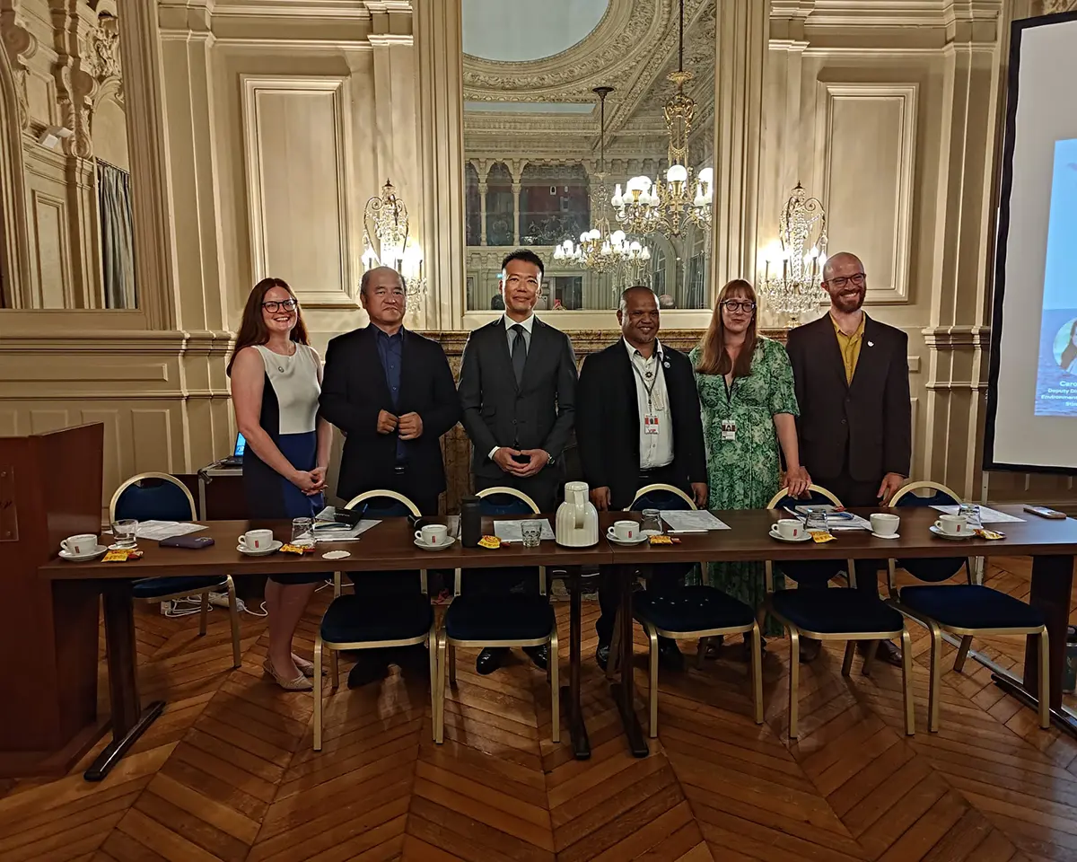 Sept personnes se tiennent debout et sourient derrière une table de conférence garnie de tasses à café et de papiers, dans une salle élégante et ornée de lustres, de parquets et d'un grand miroir reflétant le plafond.