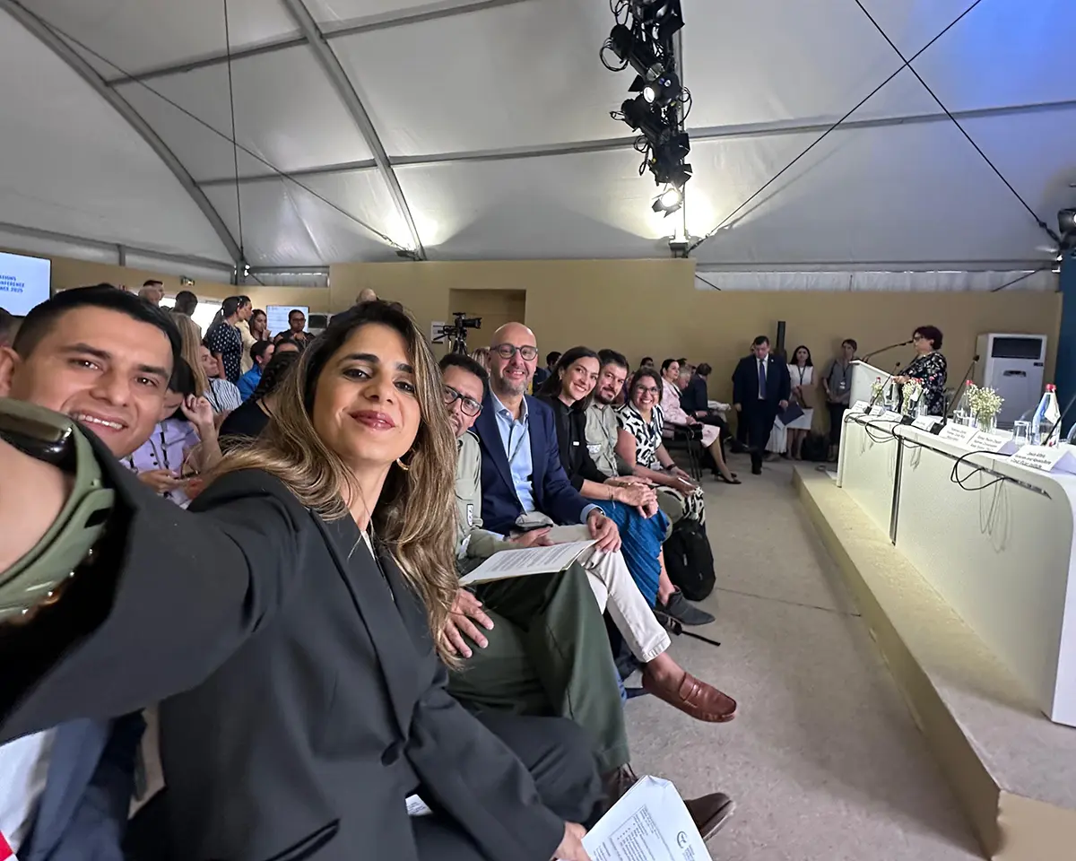 Un groupe de personnes en tenue de travail est assis dans une salle de conférence sous tente, souriant à l'appareil photo tandis que l'une d'entre elles prend un selfie. D'autres personnes sont assises ou debout à l'arrière-plan, écoutant les orateurs d'une table ronde.
