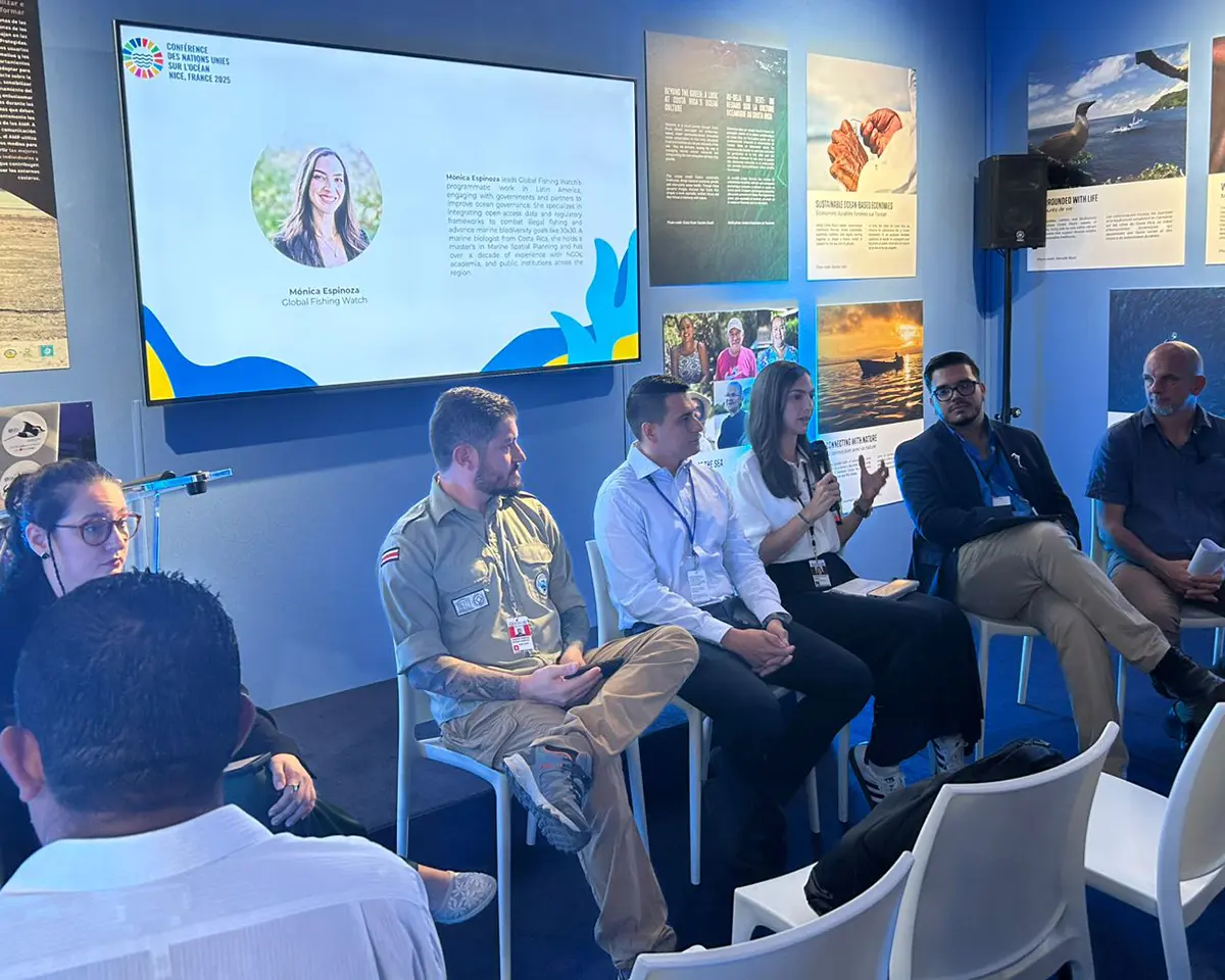 Un groupe de six personnes assises sur des chaises participe à une table ronde dans une salle aux murs bleus où se trouvent des affiches d'information et un écran affichant la photo et le texte d'une femme. Une femme parle tandis que les autres écoutent attentivement.