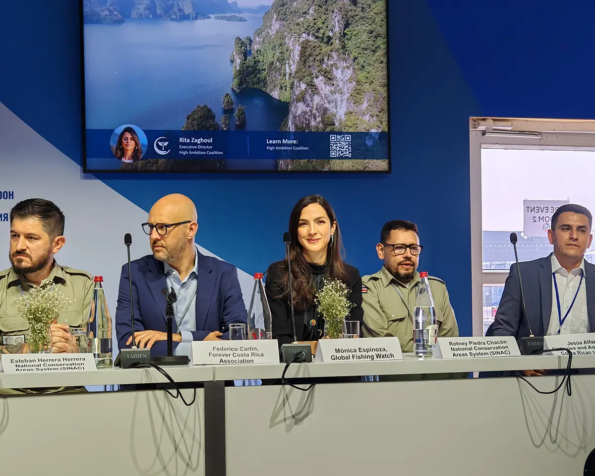Cinq personnes sont assises à une table avec des microphones pendant une table ronde. Un écran situé derrière eux affiche une image scénique, du texte et la photo d'une femme. Des plaques nominatives sont visibles devant chaque participant.