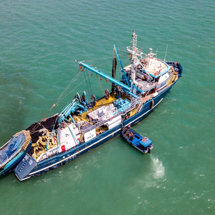 Vista aérea de un gran barco pesquero azul con el equipo en cubierta y una embarcación más pequeña amarrada a su lado en el océano verde.