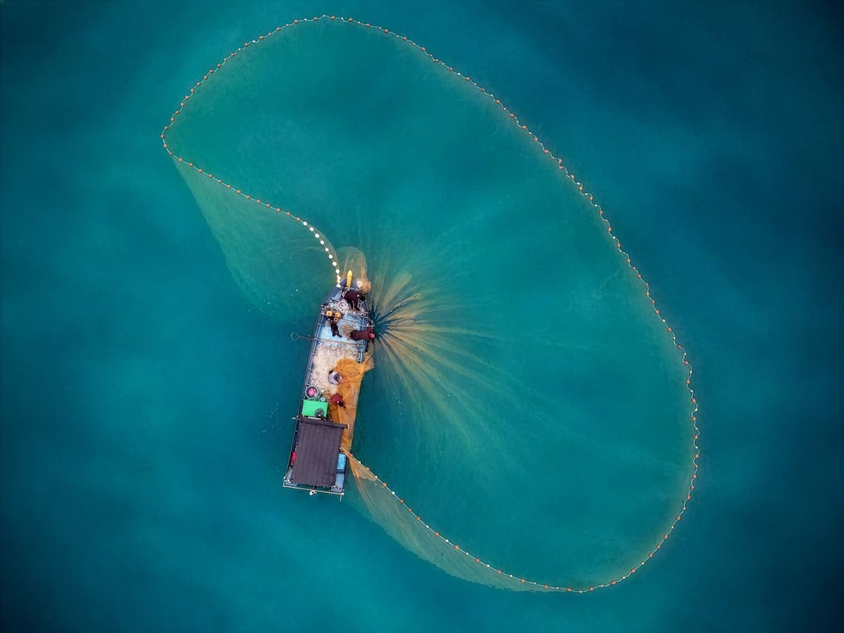 Un barco pesquero lanza una gran red circular a las aguas azules y cristalinas, creando un amplio arco que se aprecia desde arriba.